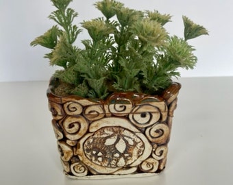 May include: A square ceramic planter with a brown and cream swirl design, filled with green and yellow artificial plants. The planter has a textured surface and a decorative, carved design on the front.