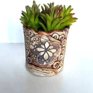 May include: A small, hand-crafted ceramic planter with a brown and white glaze. The planter features a swirling design and a raised flower in the center. The planter is filled with green succulent plants.