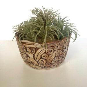 May include: A brown ceramic planter with a raised design of a person, flowers, and swirls. The planter is filled with green air plants.