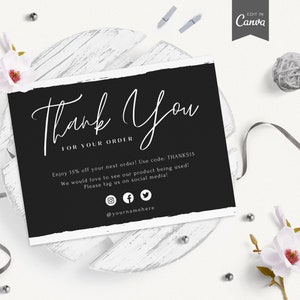 以下が含まれることがあります： 黒と白のサンキューカード。手書きのサンキューメッセージと、次回の注文で15%割引になるクーポンコードが記載されています。ソーシャルメディアのアイコンと「@yournamehere」というテキストも含まれています。