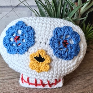 May include: White crocheted skull with blue and yellow flower accents. The skull has a red stitched mouth.