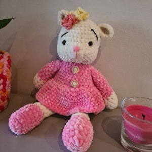 May include: A crocheted plush toy in the shape of a white lamb wearing a pink dress with white trim. The lamb has black eyes and a pink nose. The lamb is sitting on a table with a pink candle in the background.