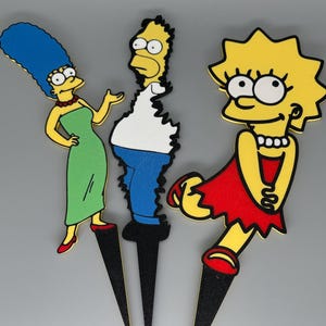 May include: Three cartoon character cutouts: Marge Simpson in a green dress, Homer Simpson in a white shirt and blue pants, and Lisa Simpson in a red dress. Each character is mounted on a black stake.