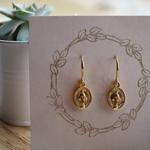 May include: Gold-tone earrings with a delicate design featuring a small oval pendant with a religious image. The earrings are displayed on a pink card with a floral design.