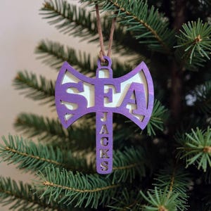 May include: A purple wooden axe-shaped Christmas ornament with the letters "SEA JACKS" cut out. The ornament hangs from a brown string against a backdrop of green pine needles.