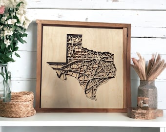 May include: A wooden wall art piece featuring a detailed laser-cut map of the state of Texas. The map is framed in a dark wood frame and is mounted on a white surface.
