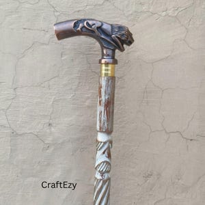 May include: A decorative walking cane with a lion's head handle. The handle is bronze-colored, and the shaft is white and brown with carved details. The cane has a gold-colored band near the top. The text "CraftEzy" is visible.