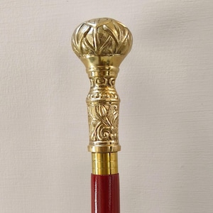 Puede incluir: Bastón decorativo con un mango dorado y ornamentado y un eje de madera rojo. El mango presenta intrincados diseños florales y una parte superior redondeada. El bastón está diseñado para brindar apoyo y estilo.