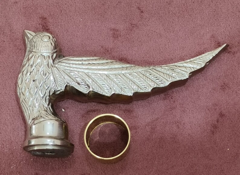 May include: Silver-toned metal bird-shaped walking stick handle with a detailed design. The handle is attached to a gold-colored metal ring.