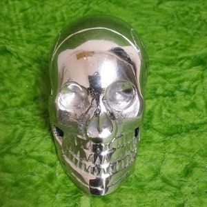May include: A silver skull sculpture with a detailed face and teeth. The skull is shiny and reflective.