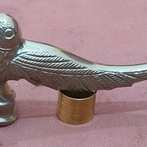 May include: A silver metal walking stick handle shaped like a bird with detailed wings. The handle is attached to a gold metal base.