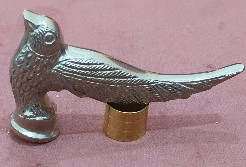 May include: A silver metal walking stick handle shaped like a bird with detailed wings. The handle is attached to a gold metal base.