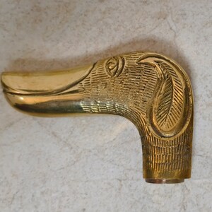 May include: A gold-toned metal duck head walking stick handle with intricate details. The handle is shaped like a duck's head with a long beak and a detailed eye.