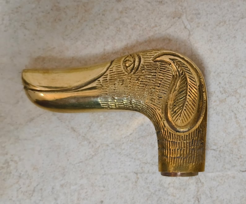May include: A gold-toned metal duck head walking stick handle with intricate details. The handle is shaped like a duck's head with a long beak and a detailed eye.