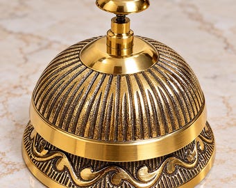 May include: A gold-colored metal service bell with a polished finish. The dome features vertical ribbed textures, while the base is decorated with an embossed wave pattern. A small, smooth, rounded knob sits at the very top of the bell.