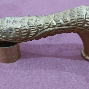 May include: A gold-colored metal cane handle with a detailed, textured design resembling a snake or dragon. The handle is curved and has a round base.