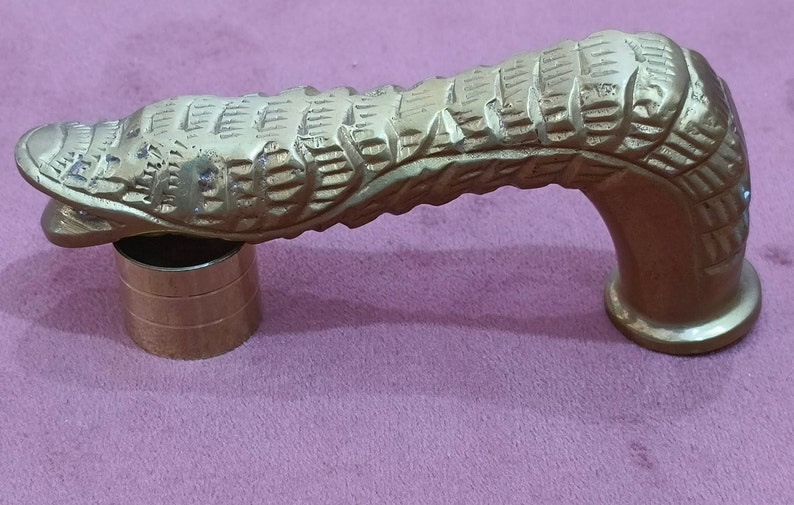 May include: A gold-colored metal cane handle with a detailed, textured design resembling a snake or dragon. The handle is curved and has a round base.
