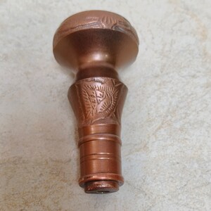 May include: A copper-coloured metal bottle stopper with an intricate design. The stopper has a round top and a cylindrical base.