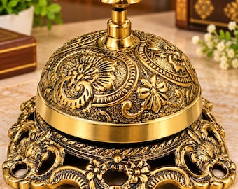 Puede incluir: Un timbre de servicio de metal dorado con un patrón floral y de volutas en relieve en la cúpula y una base ancha y decorativa con detalles recortados. El timbre tiene un botón superior liso y pulido para presionar.