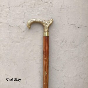 Op de afbeelding: Een houten wandelstok met een messing handvat. Het handvat heeft een sierlijk, gebogen ontwerp. De schacht is roodbruin van kleur met een paar messing accenten. De stok is ongeveer 91 cm lang.