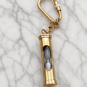 May include: A gold-colored hourglass keychain. The hourglass is encased in a cylindrical brass tube with a clear window, allowing a view of the sand. The keychain includes a clasp and chain.