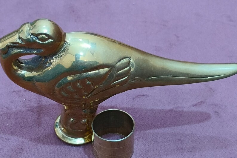 May include: A gold-colored metal bird-shaped walking stick handle with intricate details. The handle is attached to a round metal base.