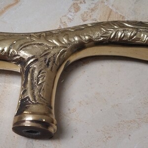 May include: A gold-colored metal cane handle with an ornate, floral design. The handle is shaped like a curved 'T' and has a smooth, polished finish.
