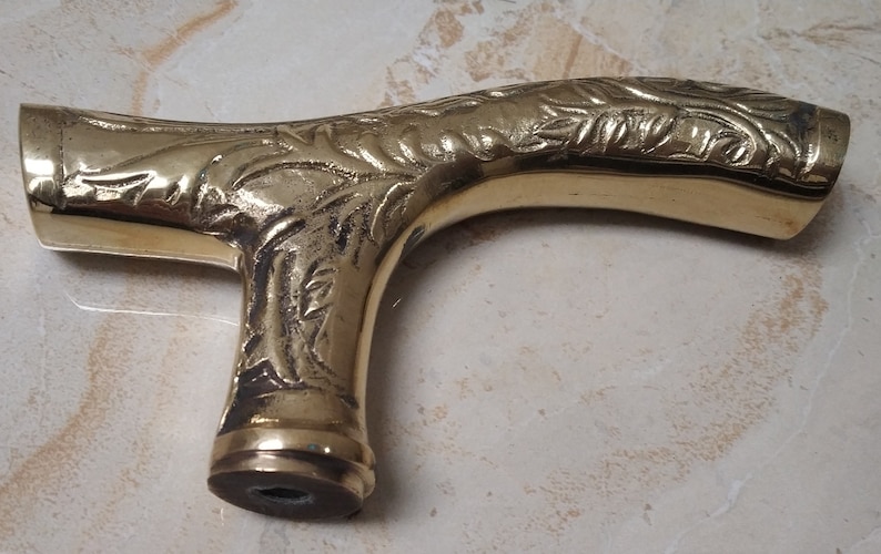 May include: A gold-colored metal cane handle with an ornate, floral design. The handle is shaped like a curved 'T' and has a smooth, polished finish.