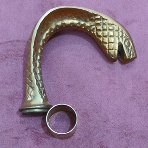 May include: A gold-colored metal cane handle shaped like a snake's head with a textured surface. The handle is attached to a round metal ring.