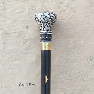 Peut inclure: Une canne noire avec une poignée décorative. La poignée est blanche avec un motif tourbillonnant noir. La canne a une bande dorée et un emblème doré. Le texte "CraftEzy" est visible.