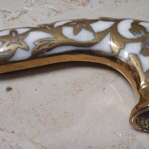 May include: A brass cane handle with a white enamel floral design. The handle is curved and has a threaded hole at the bottom.