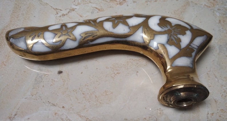 May include: A brass cane handle with a white enamel floral design. The handle is curved and has a threaded hole at the bottom.