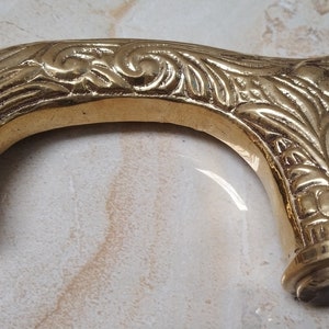 May include: A gold-colored metal walking stick handle with an ornate floral design. The handle is curved and has a smooth, polished finish.