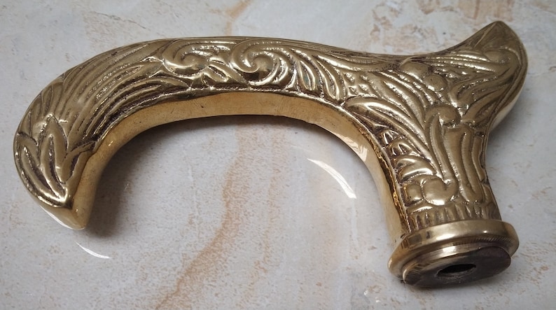 May include: A gold-colored metal walking stick handle with an ornate floral design. The handle is curved and has a smooth, polished finish.