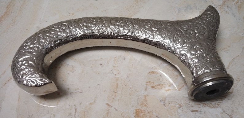 May include: Silver-toned walking stick handle with an ornate floral design. The handle is curved and has a round base.