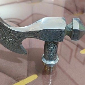 May include: A silver-coloured metal axe head with intricate floral designs. The axe head is attached to a wooden handle.