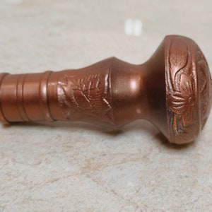 May include: A copper-coloured metal bottle stopper with a decorative floral design. The stopper has a cylindrical base and a rounded top.