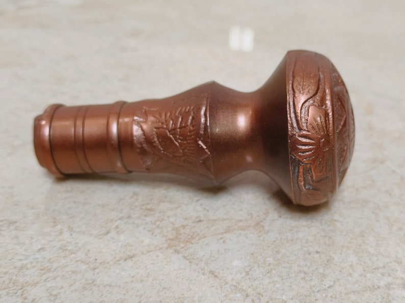 May include: A copper-coloured metal bottle stopper with a decorative floral design. The stopper has a cylindrical base and a rounded top.