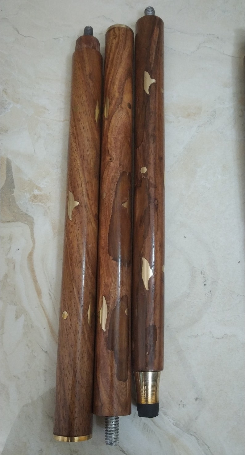 Op de afbeelding: Drie cilindrische houten wandelstoksegmenten met een gepolijste bruine nerf en ingelegde koperen accenten in blad- en puntvormen. De segmenten bevatten metalen schroefverbindingen aan de uiteinden en een zwarte rubberen dop aan de onderkant.