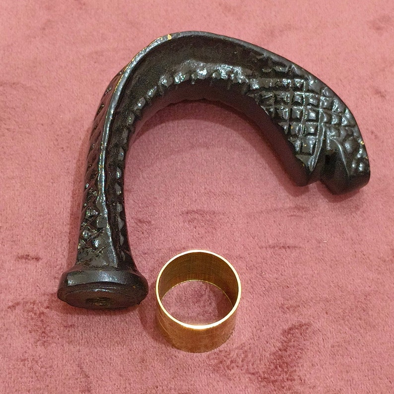 May include: A black, intricately carved, wooden cane handle with a decorative pattern. The handle is shaped like a curved hook and has a gold metal ring attached to the bottom.