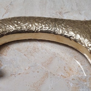 May include: A gold-colored metal cane handle with an intricate floral design. The handle is curved and has a round base with a small hole in the center.