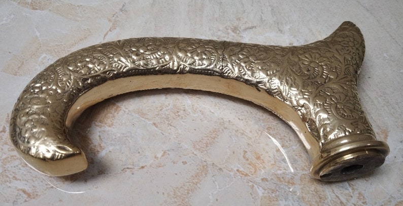 May include: A gold-colored metal cane handle with an intricate floral design. The handle is curved and has a round base with a small hole in the center.