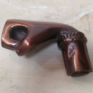 May include: A bronze-coloured cane handle shaped like a hand making a fist. The handle has a smooth, polished finish with a detailed design at the wrist. The bottom of the handle is cylindrical.
