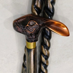 May include: A walking stick with a detailed bronze rabbit head handle. The handle has a copper colored ear and a brass collar. The stick is made of dark wood with a twisted pattern.