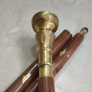 May include: A wooden walking stick with a brass top. The top features a decorative design of leaves and a ring around the base. The stick is made of dark wood and has a smooth finish.
