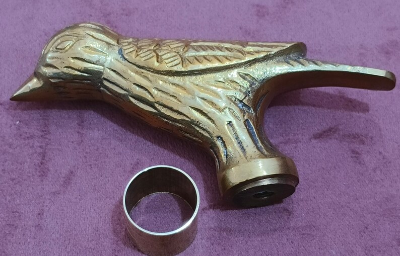 May include: A gold-colored metal bird-shaped cane handle with a decorative design. The handle has a round base with a small hole in the center. A silver-colored metal ring is also included.