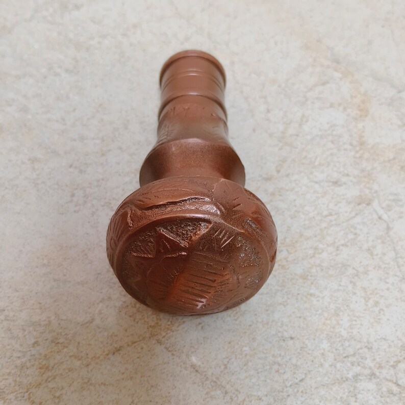 May include: A copper bottle stopper with an intricate carved design. The stopper is shaped like a rounded top with a cylindrical neck.