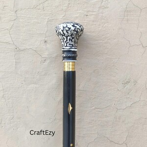 May include: A black walking cane with a decorative, patterned white and black top. The cane features a gold-coloured band and a gold-coloured accent near the middle. The text "CraftEzy" is visible near the bottom.