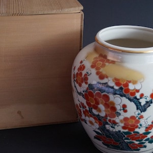 May include: A white ceramic vase with a gold rim and a floral design featuring red, orange, and white flowers on a black branch. The vase is sitting on a wooden box.