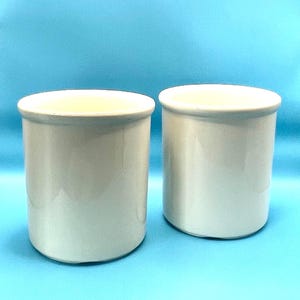 Może przedstawiać: Dwa białe ceramiczne kubki lub pojemniki. Kubki mają cylindryczny kształt i gładkie, błyszczące wykończenie.